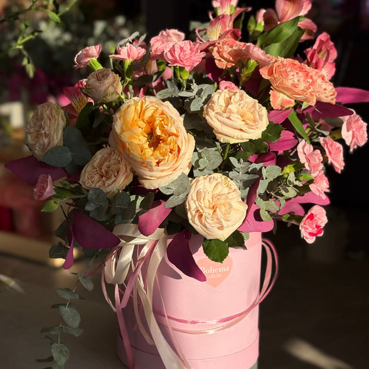 Rožinių pastelinių spalvų gėlių kompozicija dėžutėje su švelniais rožiniais, persikiniais ir kreminiais atspalviais, apvali ir minkštos struktūros forma